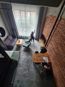 Photo taken from loft section of apartment looking down at room with woman waving at camera