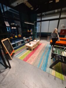 Area for kids' with little tables and chairs, fussball and toys with colourful rug. Woman facing away from camera.