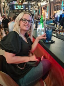 Girl sitting at bar with a blue cocktail, that has a plastic shark on top of it.